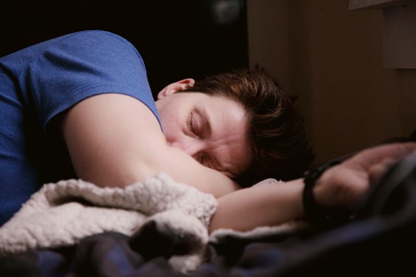 Photograph of a man lying down on a bed on his side asleep, his right hand is resting under his head.