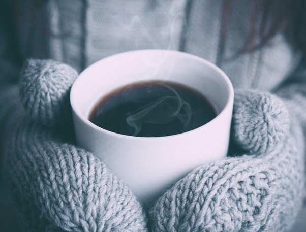 Picture of a person wearing wooly grey mittens holding a hot cup of coffee with steam coming off it