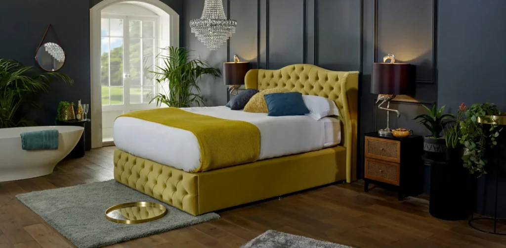 Elegant bedroom with a yellow upholstered bed, dark walls, and wooden floors. A chandelier, plants, and a large window enhance the space.